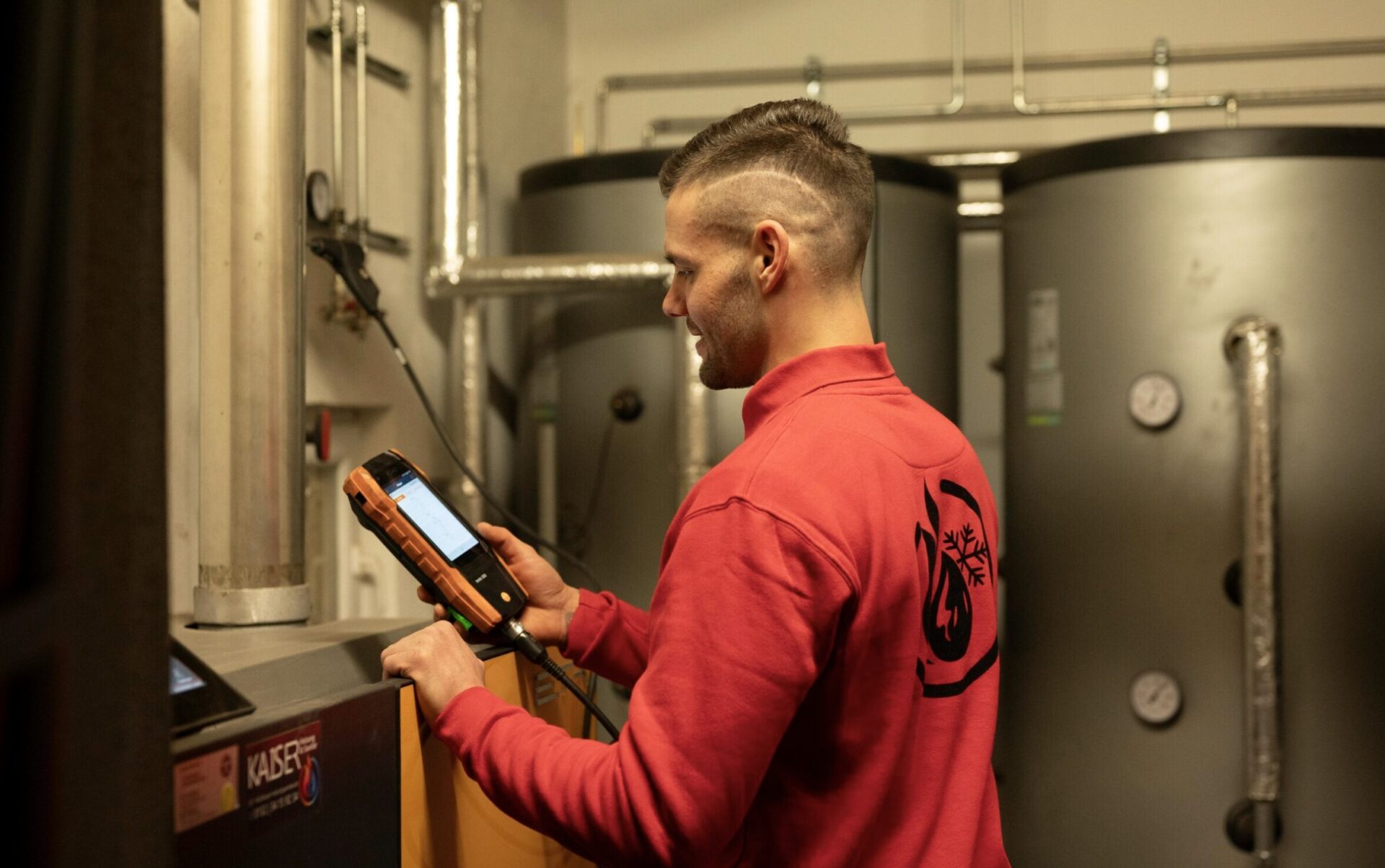 Techniker in rotem Pullover, der ein Messgerät in einem Heizungsraum bedient.