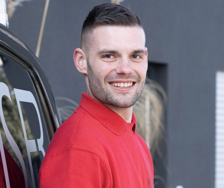 Mann mit kurzem Haar in einem roten Poloshirt lächelt vor einem Fahrzeug mit sichtbarem Logo.